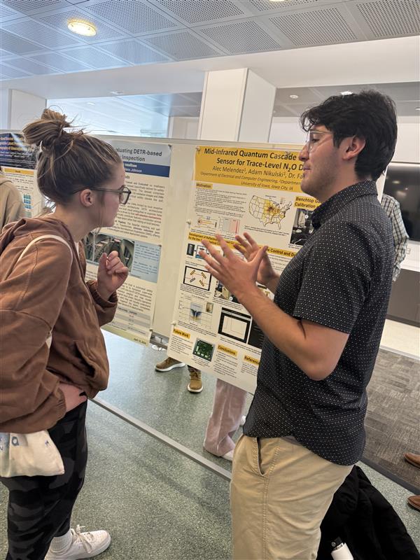 Two people stand in front of a research poster in a public indoor space, facing each other and gesturing with their hands while discussing the poster’s content during a research presentation session.