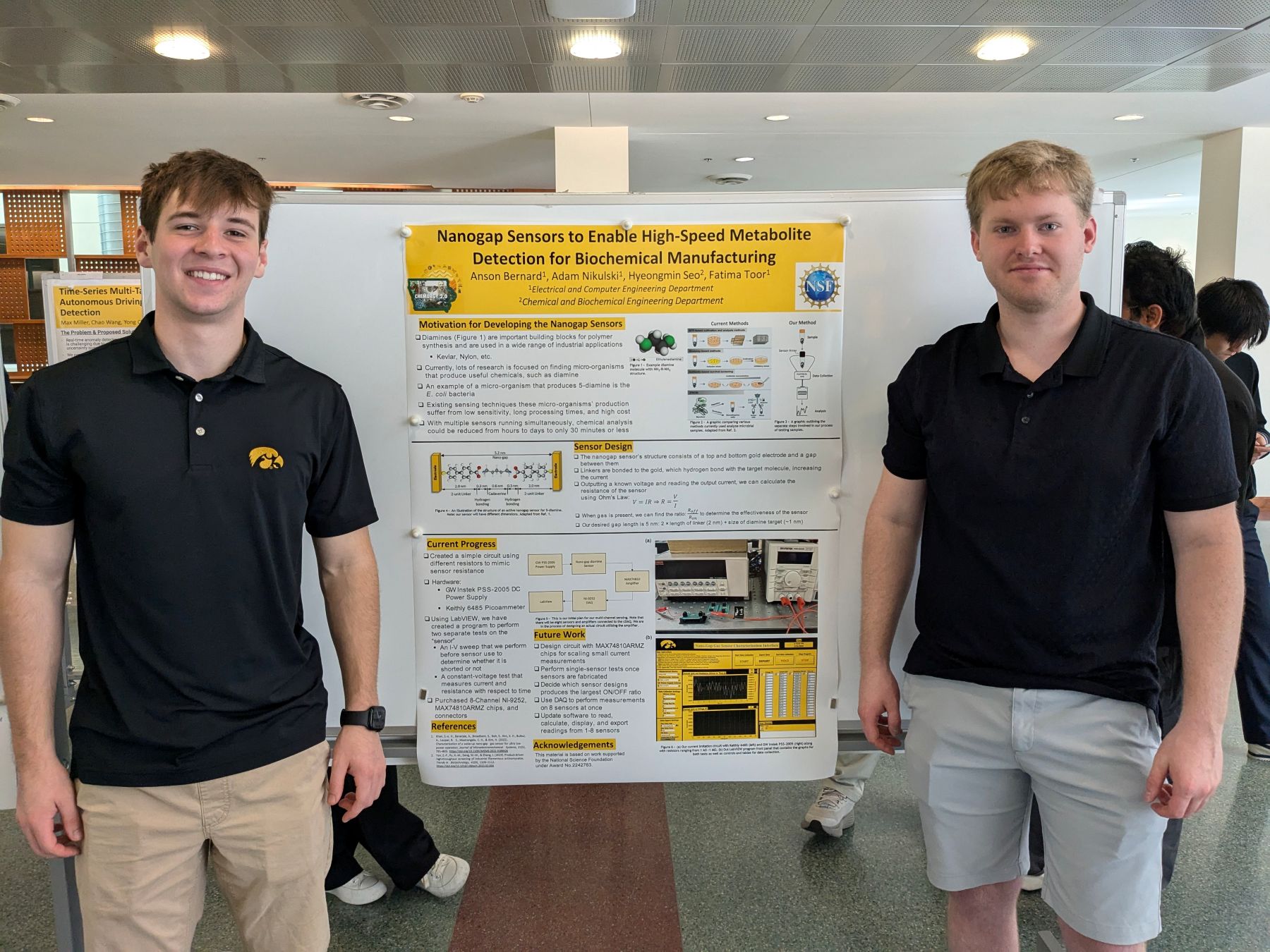 Two undergraduate researchers stand on either side of a scientific poster titled about nanoscale sensors, displayed on a freestanding poster board in a university building.