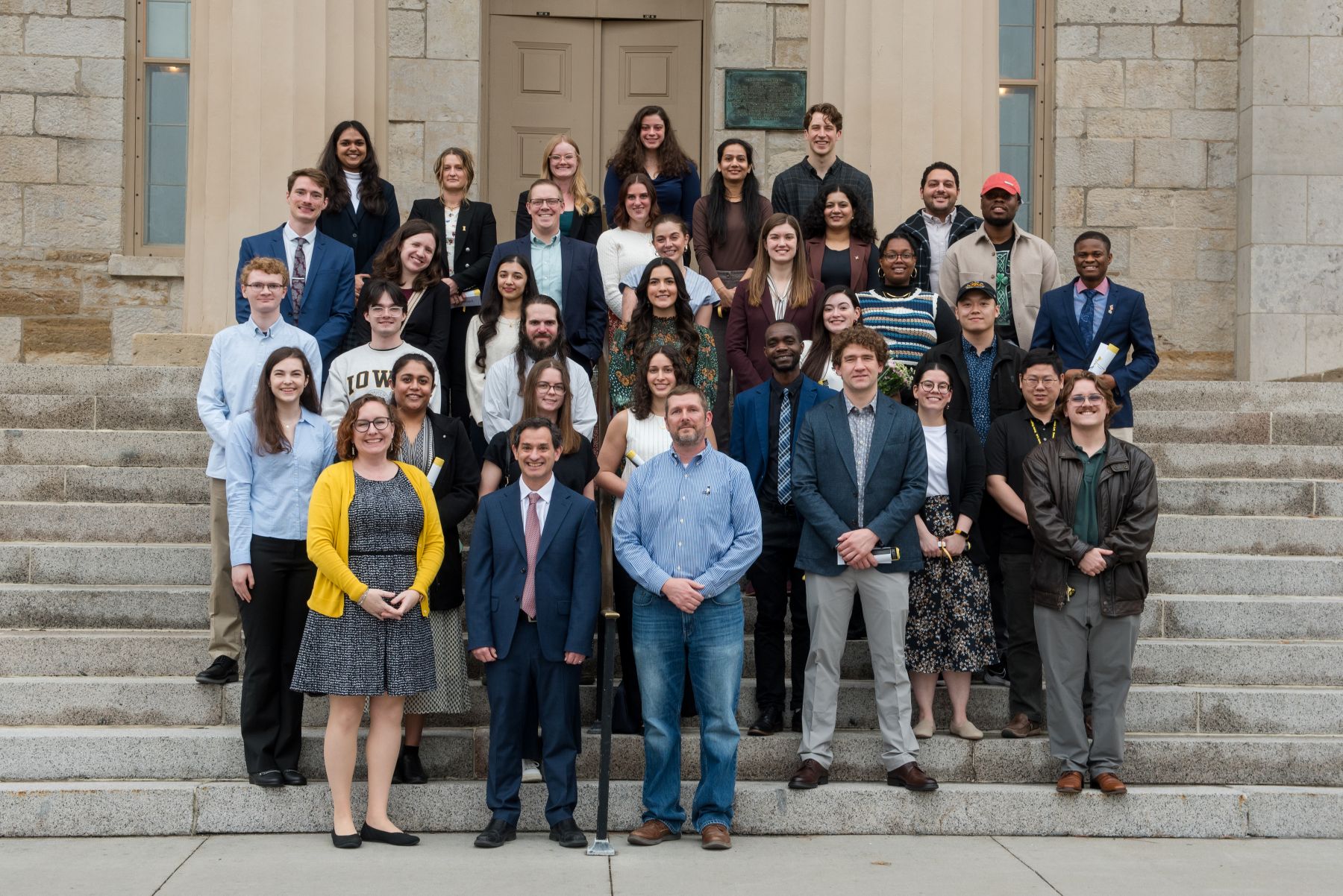 A large group of people pose together on stone steps outside a university building.